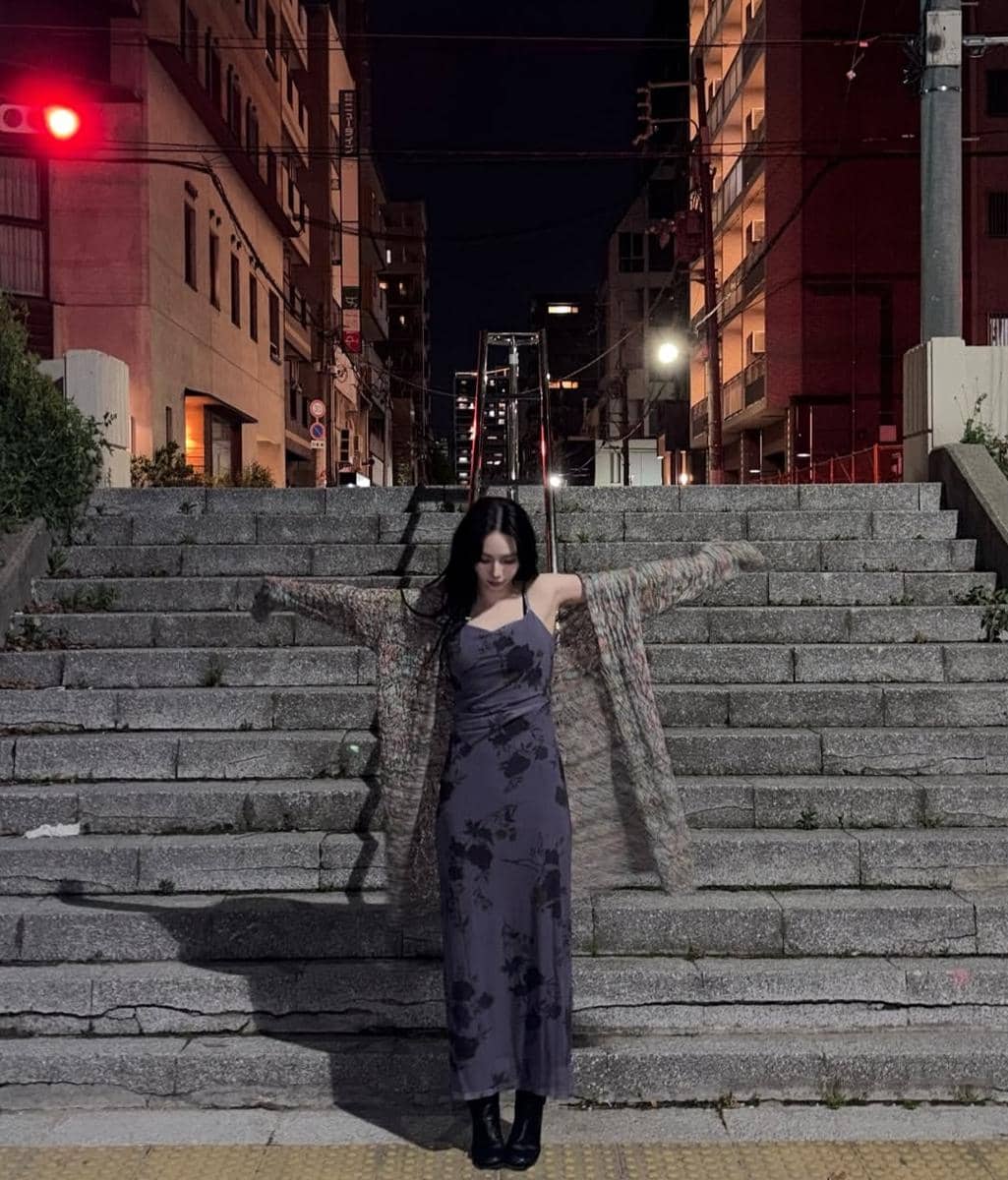 Seorang wanita berambut panjang mengenakan gaun ungu bermotif bunga berdiri di tangga kota pada malam hari dengan lampu jalan menyala.