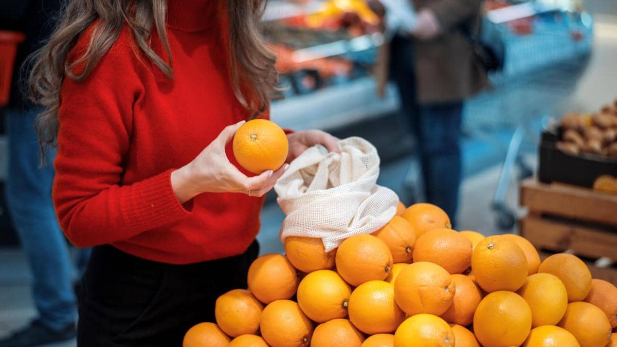 Ilustrasi seorang perempuan memilih dan membeli jeruk segar yang dipajang di bagian buah-buahan sebuah supermarket.