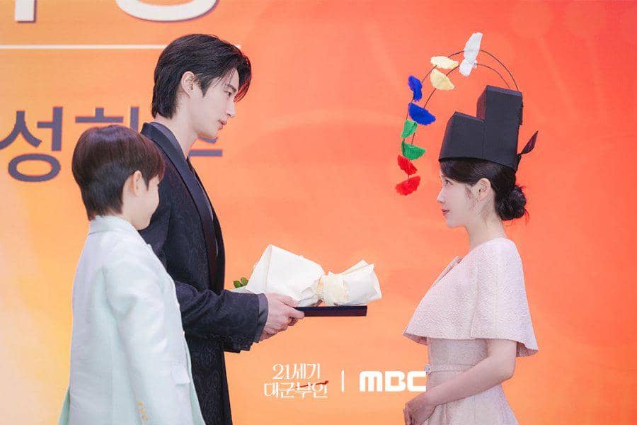 Dua orang berdiri saling berhadapan di depan latar oranye, salah satunya mengenakan topi tradisional dengan hiasan warna-warni dan menerima hadiah.