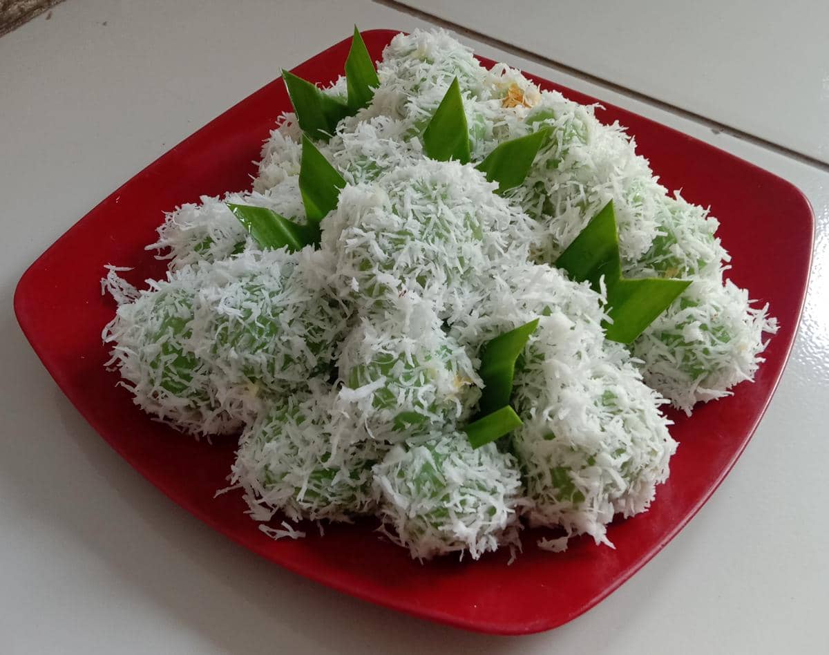 Foto produk klepon hijau di atas piring merah dengan taburan kelapa melimpah.
