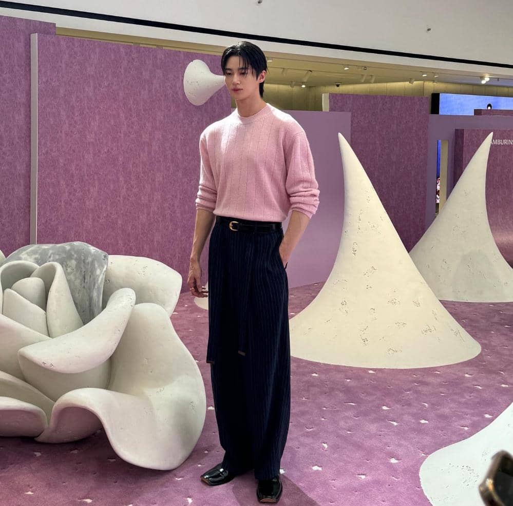 Seorang pria mengenakan sweater pink dan celana panjang hitam bergaris berdiri di ruang pameran bernuansa ungu dengan dekorasi artistik.