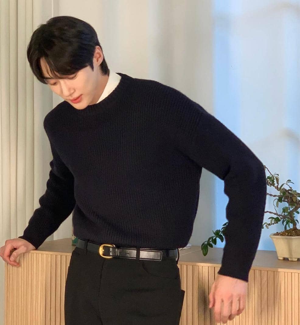 Seorang pria berambut hitam mengenakan sweater rajut hitam dan celana gelap berdiri di dekat meja kayu dengan tanaman hias kecil.
