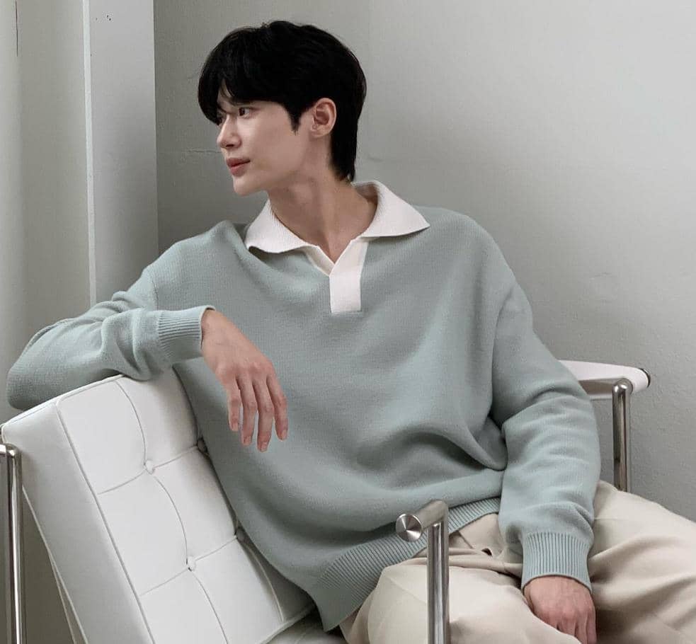 Seorang pria berambut hitam mengenakan sweater hijau muda dengan kerah putih duduk santai di kursi berwarna putih.