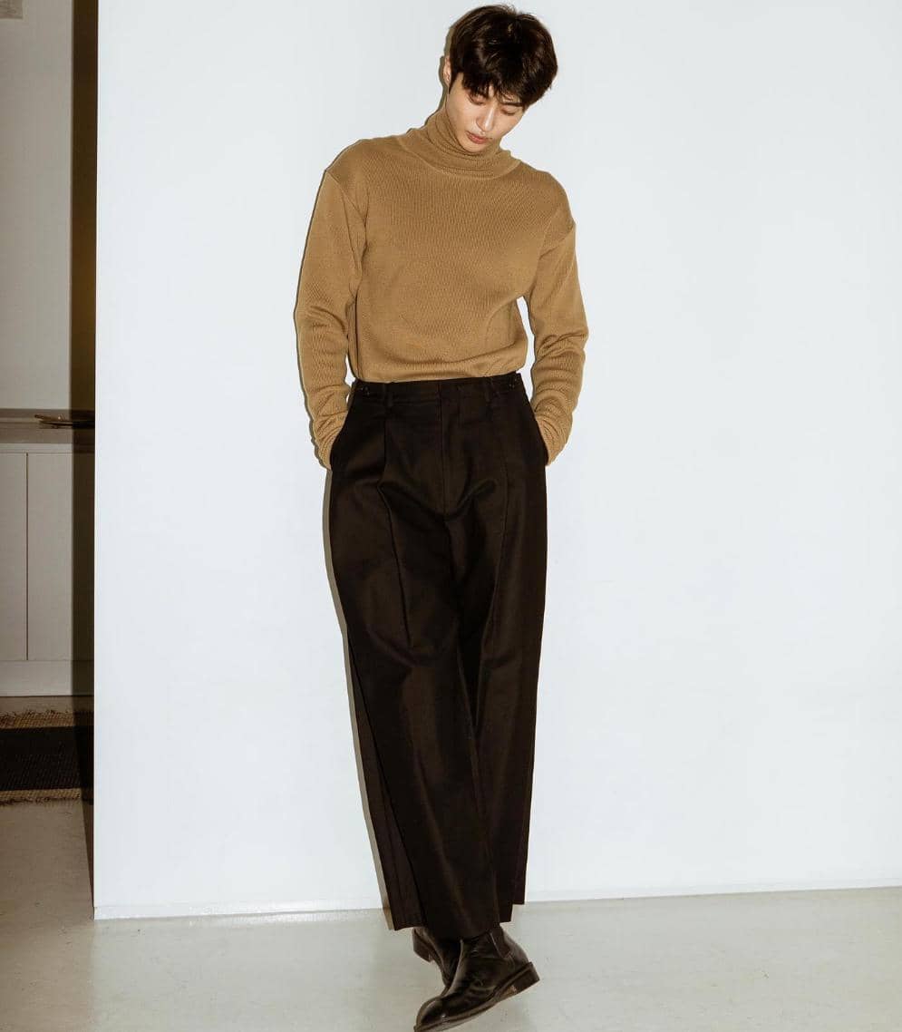 Seorang pria mengenakan sweater cokelat dan celana panjang hitam berdiri bersandar di dinding putih dengan gaya kasual elegan.