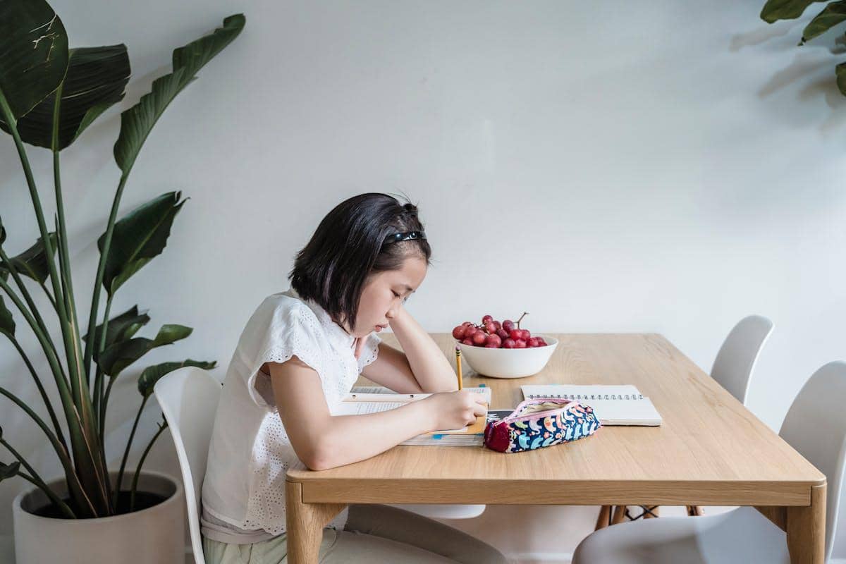 Seorang anak perempuan belajar dengan fokus di meja kayu, menulis di buku dengan pensil di ruang yang tenang dan minim distraksi.