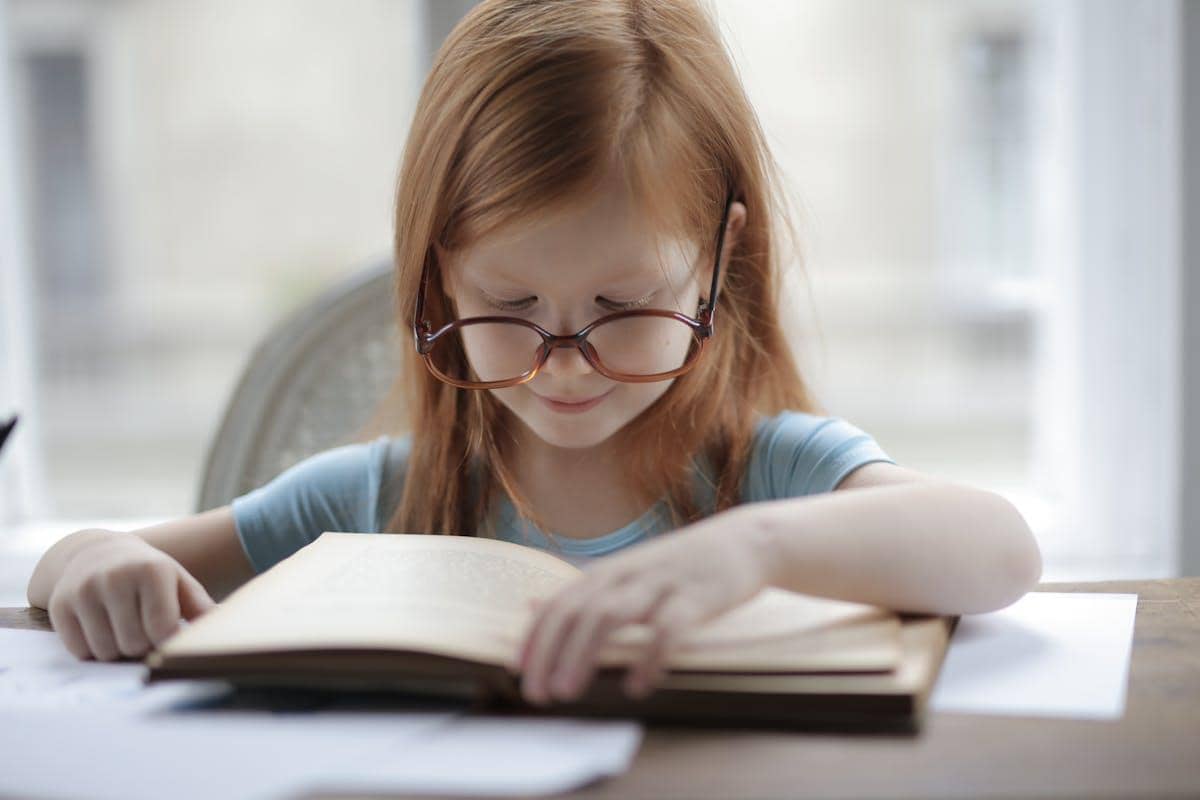 Seorang anak perempuan berambut merah mengenakan kacamata sedang membaca buku di meja dengan latar belakang jendela terang.