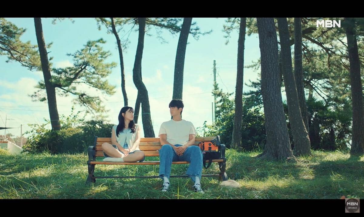 Dua orang duduk di bangku taman di bawah pepohonan rindang pada siang hari, dengan suasana tenang dan langit cerah.