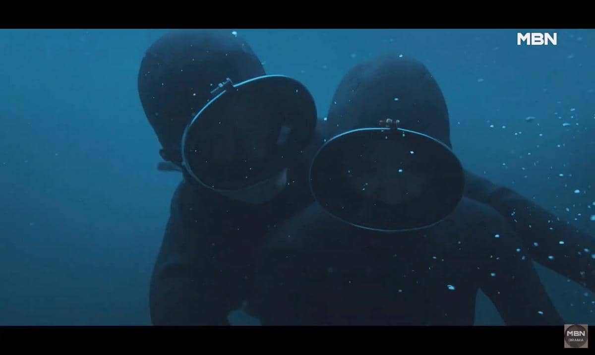 Dua penyelam mengenakan pakaian selam hitam dan masker besar sedang berada di bawah air dalam cuplikan teaser drama Korea Azure Spring.