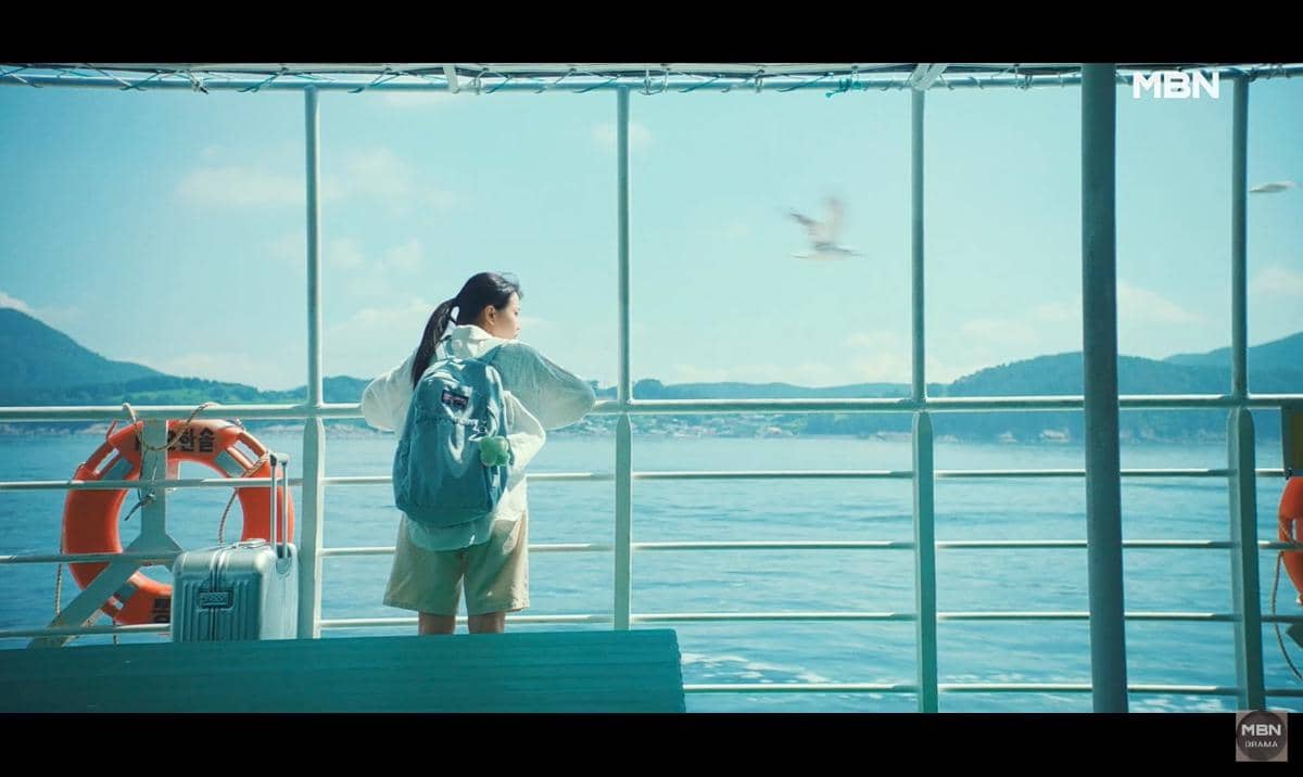 Seorang perempuan dengan ransel biru berdiri di atas kapal feri, memandangi laut biru dan pegunungan di kejauhan di bawah langit cerah.