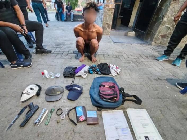 Seorang pria tanpa baju berjongkok di depan barang bukti berupa tas, topi, masker, dokumen, dan berbagai benda lainnya yang disita petugas.