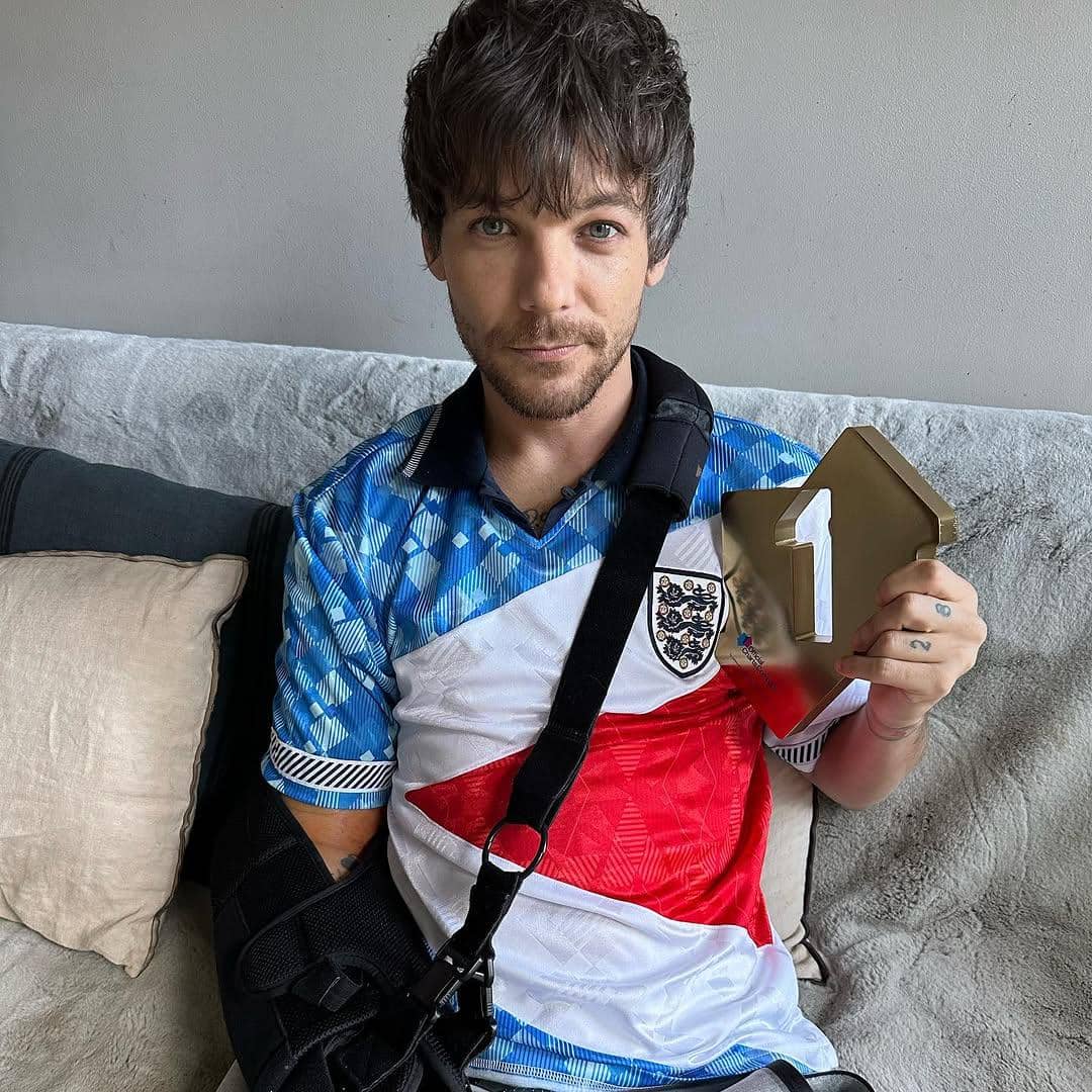 Seorang pria mengenakan jersey bergaya Inggris duduk di sofa sambil memegang trofi berbentuk angka satu berwarna emas.