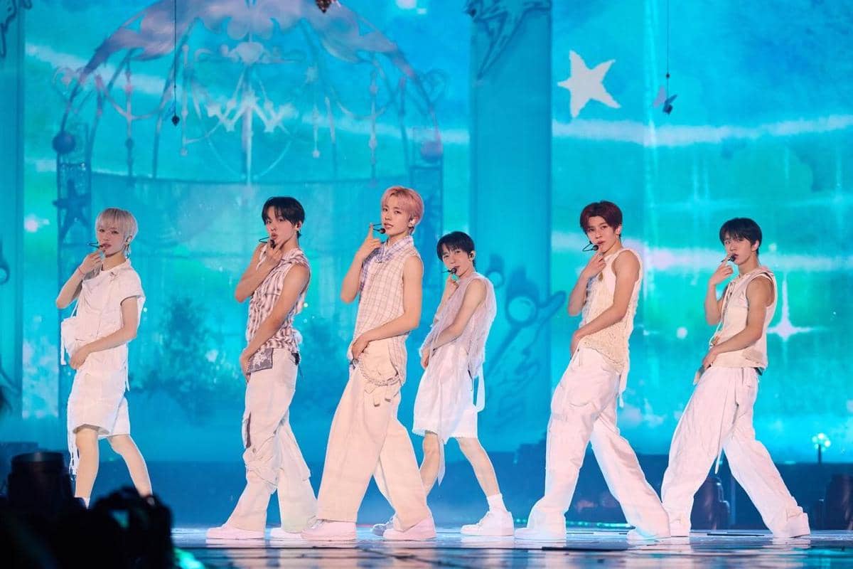 Enam anggota grup musik pria tampil di atas panggung dengan pencahayaan biru, mengenakan busana putih dan melakukan koreografi terkoordinasi.