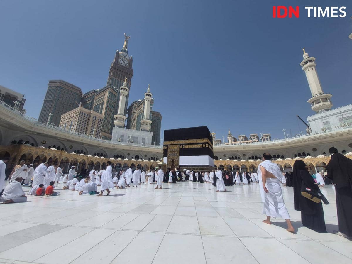 Jemaah mengenakan pakaian ihram beribadah di sekitar Ka'bah di Masjidil Haram, Makkah, dengan latar menara jam Abraj Al Bait di bawah langit cerah.