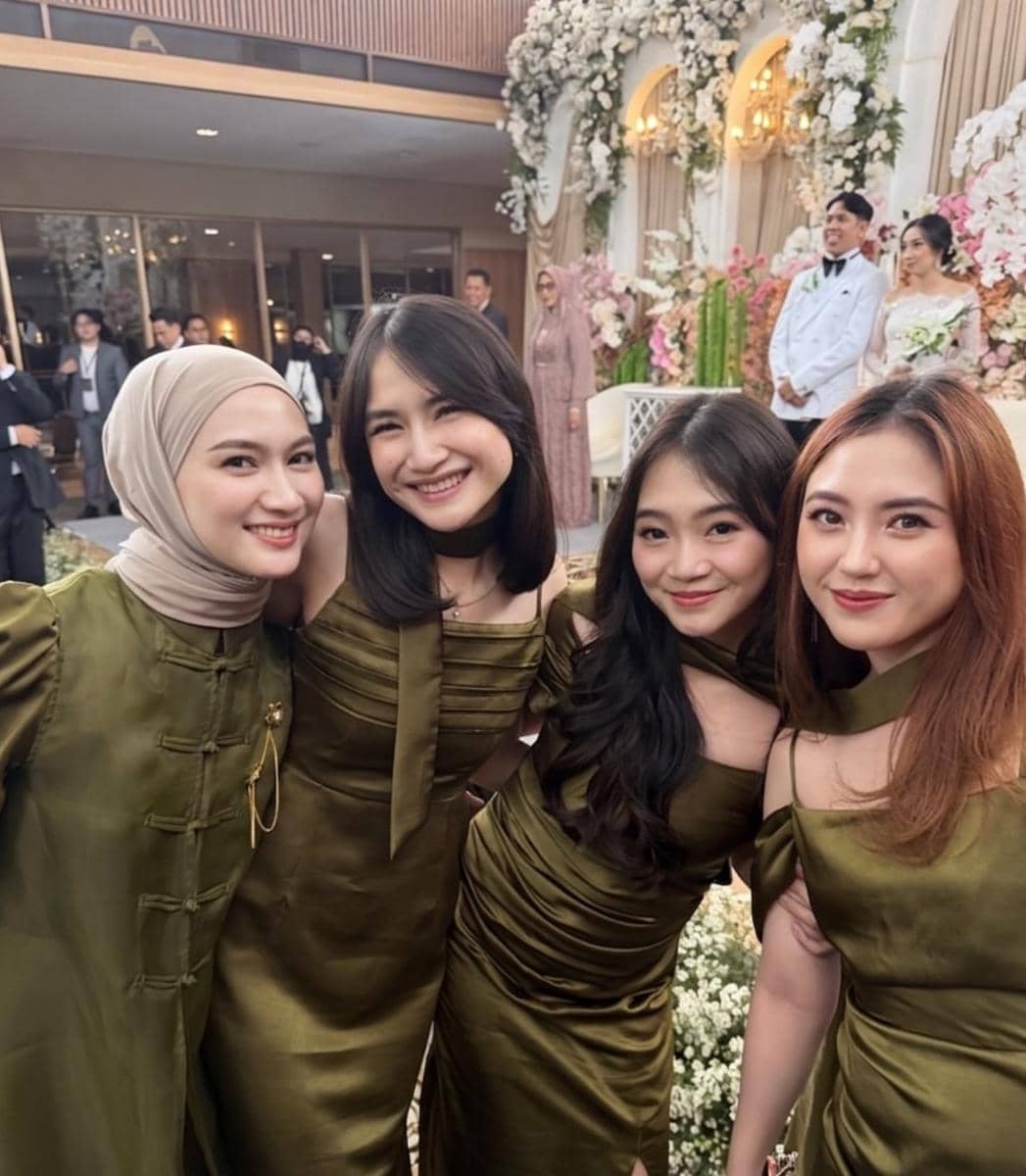 Empat wanita bergaun hijau zaitun berpose tersenyum di acara pernikahan dengan latar dekorasi bunga dan pengantin di pelaminan.