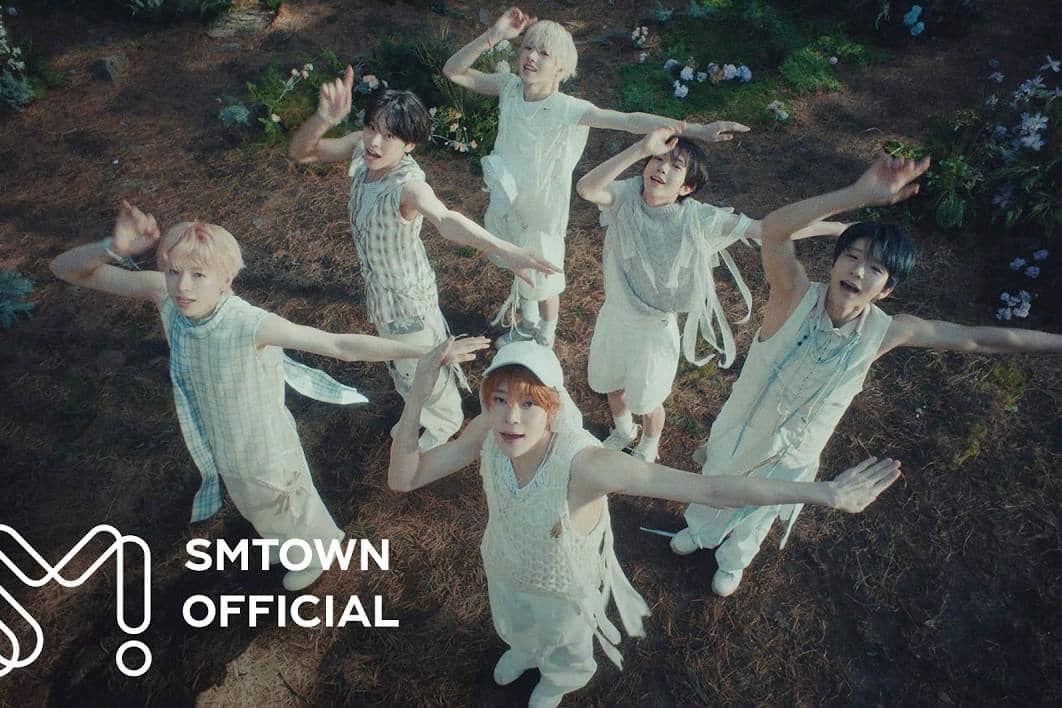 Enam anggota grup musik tampil energik di luar ruangan dengan busana putih dan latar pepohonan serta bunga berwarna lembut.