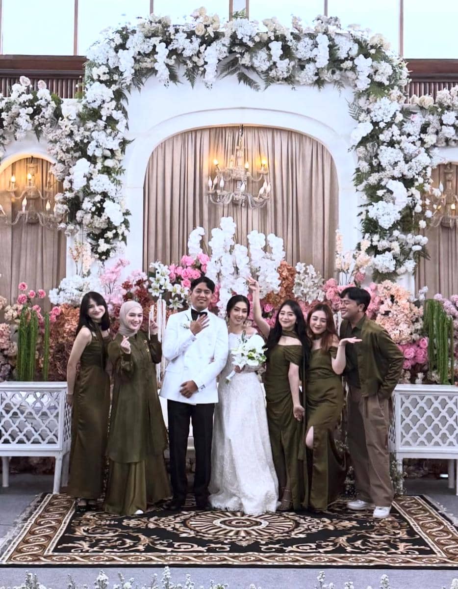 Pasangan pengantin berpose bersama teman-teman di pelaminan berhias bunga putih dan pastel dalam suasana pernikahan elegan.