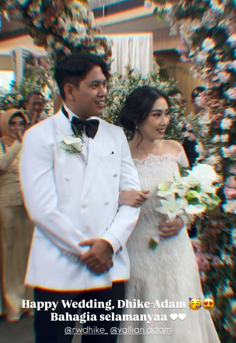Sepasang pengantin tersenyum bahagia saat berjalan bersama di acara pernikahan, dikelilingi tamu dan dekorasi bunga indah.