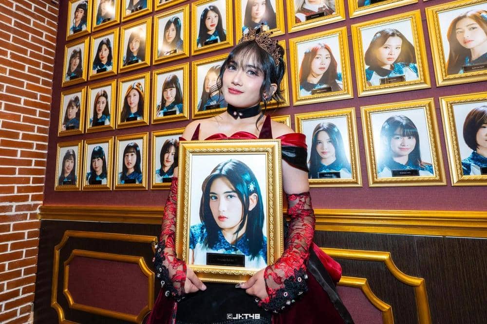 Seorang perempuan mengenakan gaun merah hitam dan mahkota memegang bingkai foto dirinya di depan dinding penuh potret berbingkai emas.