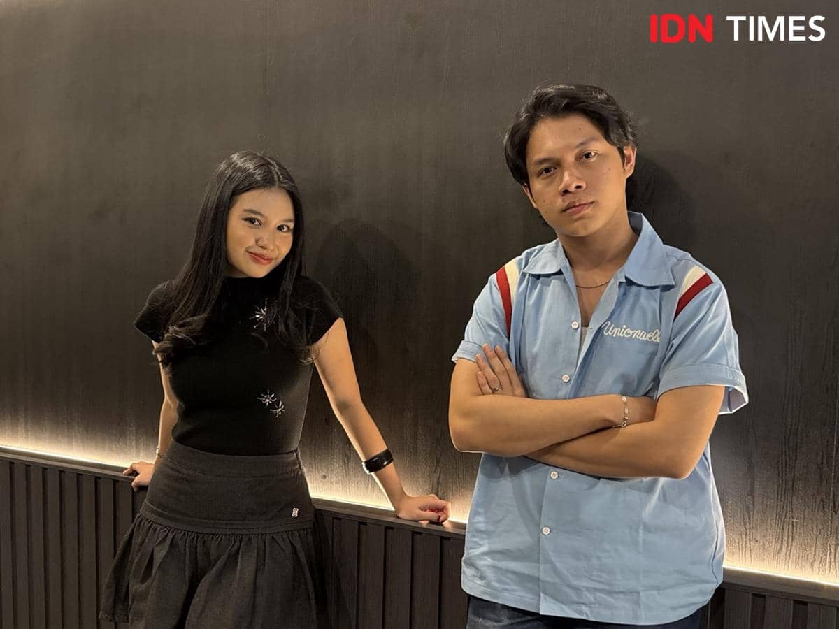 Seorang perempuan berambut panjang mengenakan atasan hitam dan rok gelap berdiri di samping seorang pria berkaus biru muda di depan dinding gelap.