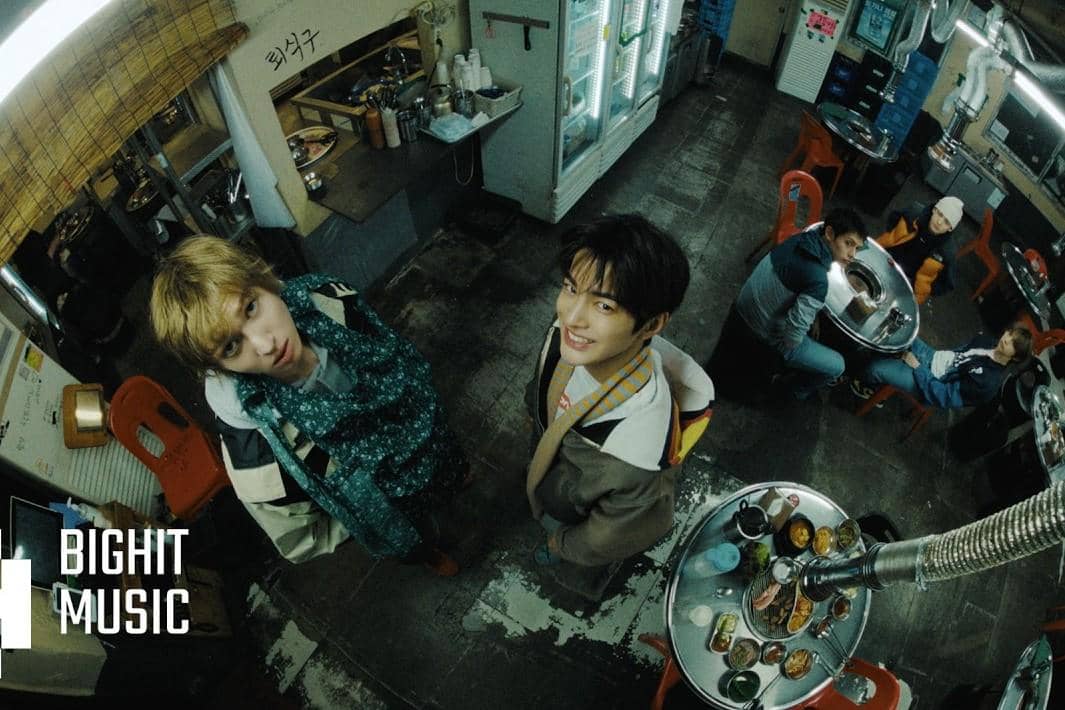 Beberapa anggota grup musik berada di restoran dengan meja bulat dan kursi merah, dua di antaranya berdiri menatap kamera.