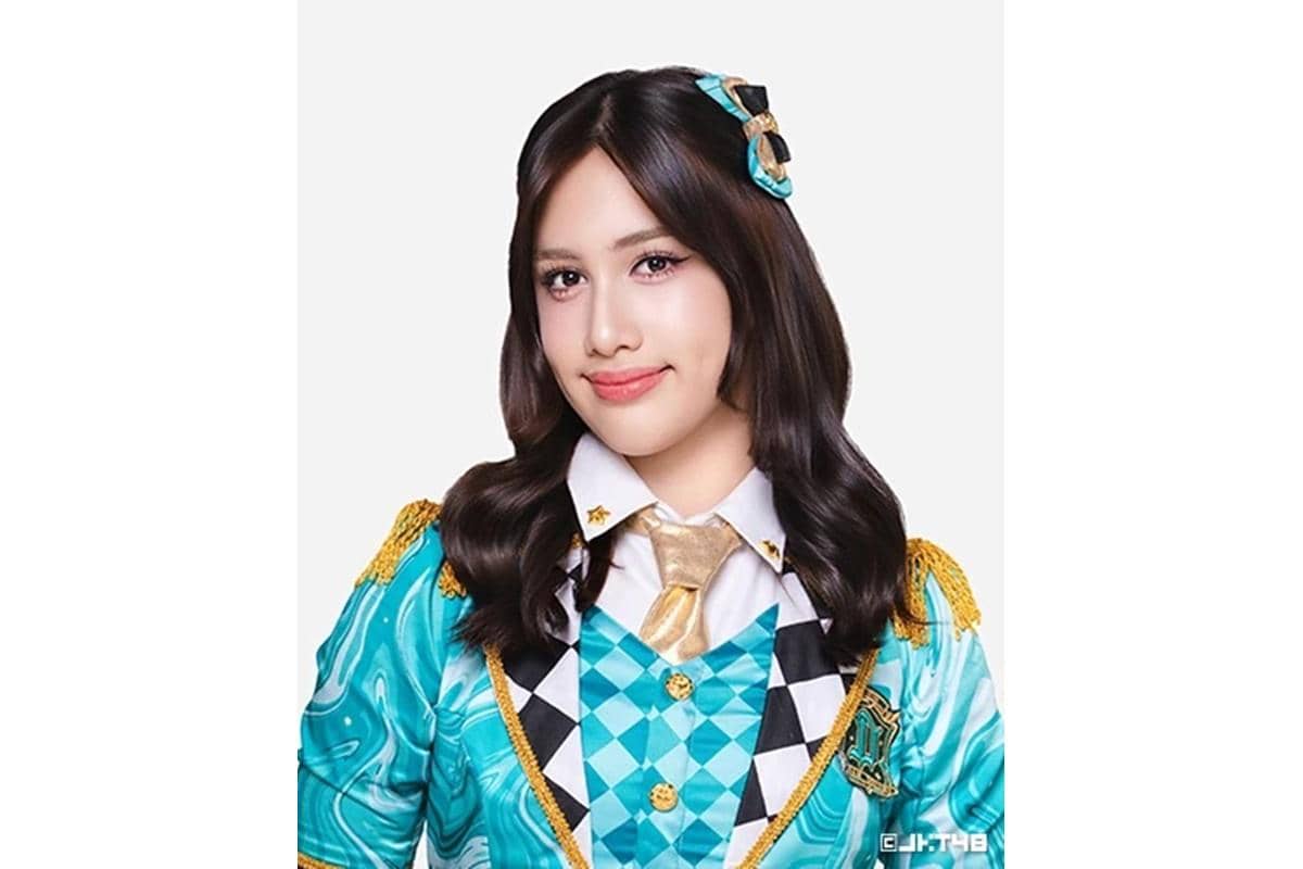 Seorang perempuan berambut hitam panjang mengenakan kostum panggung berwarna biru toska dengan dasi emas dan hiasan rambut serasi.