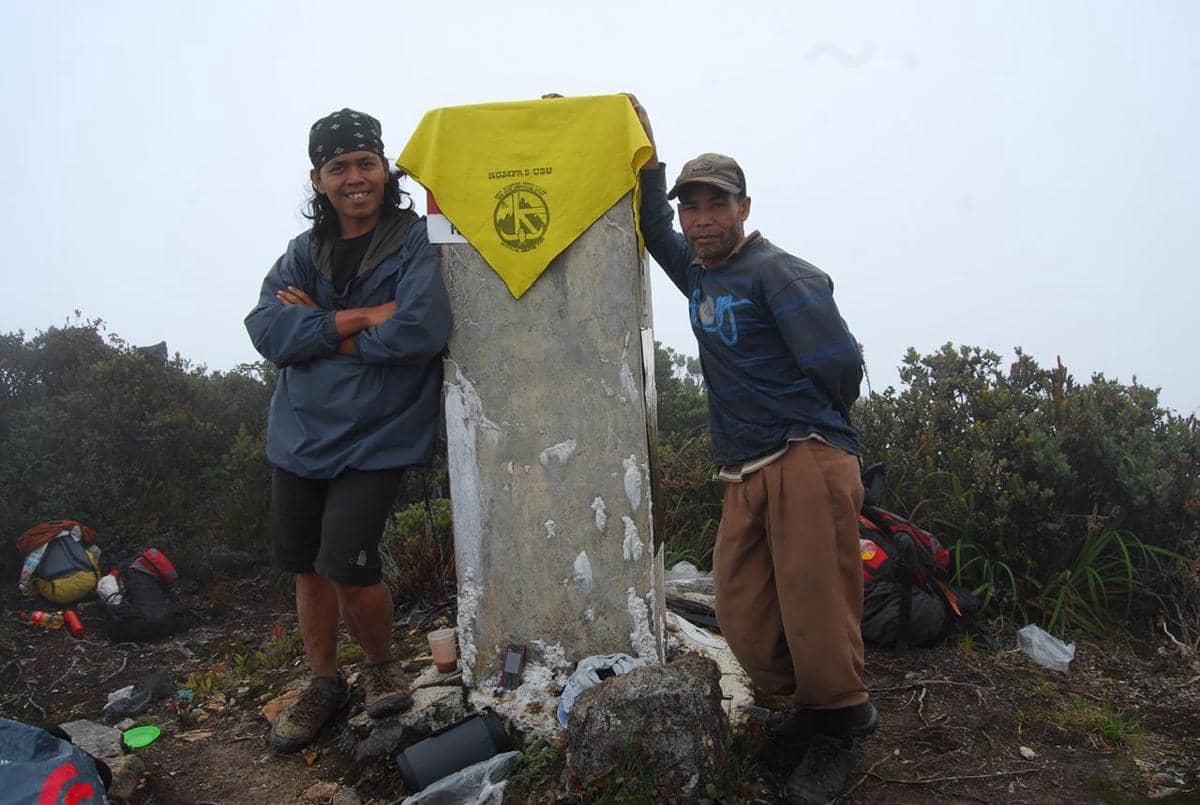 Dua pendaki berdiri di puncak Gunung Leuser dengan latar kabut tebal, bersandar pada tugu penanda puncak yang tertutup kain kuning.
