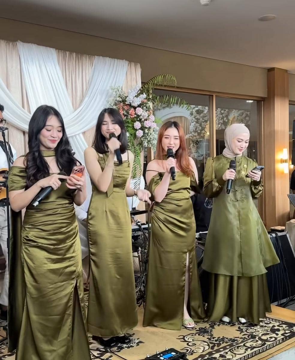 Empat wanita mengenakan gaun hijau zaitun sedang bernyanyi di acara pernikahan dengan latar dekorasi bunga dan tirai putih.