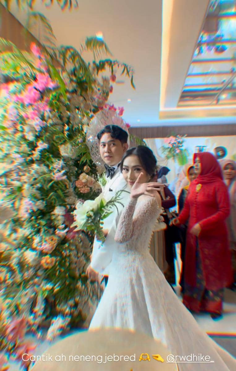 Pasangan pengantin berjalan bersama di pelaminan dengan latar bunga warna-warni, sang pengantin wanita melambaikan tangan sambil tersenyum.