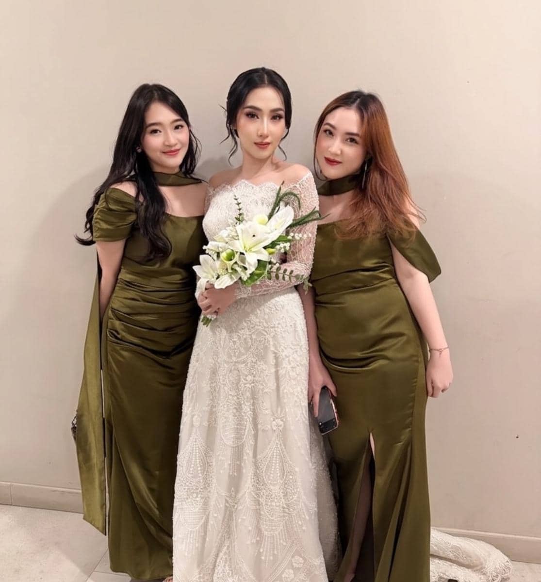 Seorang pengantin bergaun putih berpose bersama dua wanita bergaun hijau zaitun di sebuah ruangan dengan dinding krem.