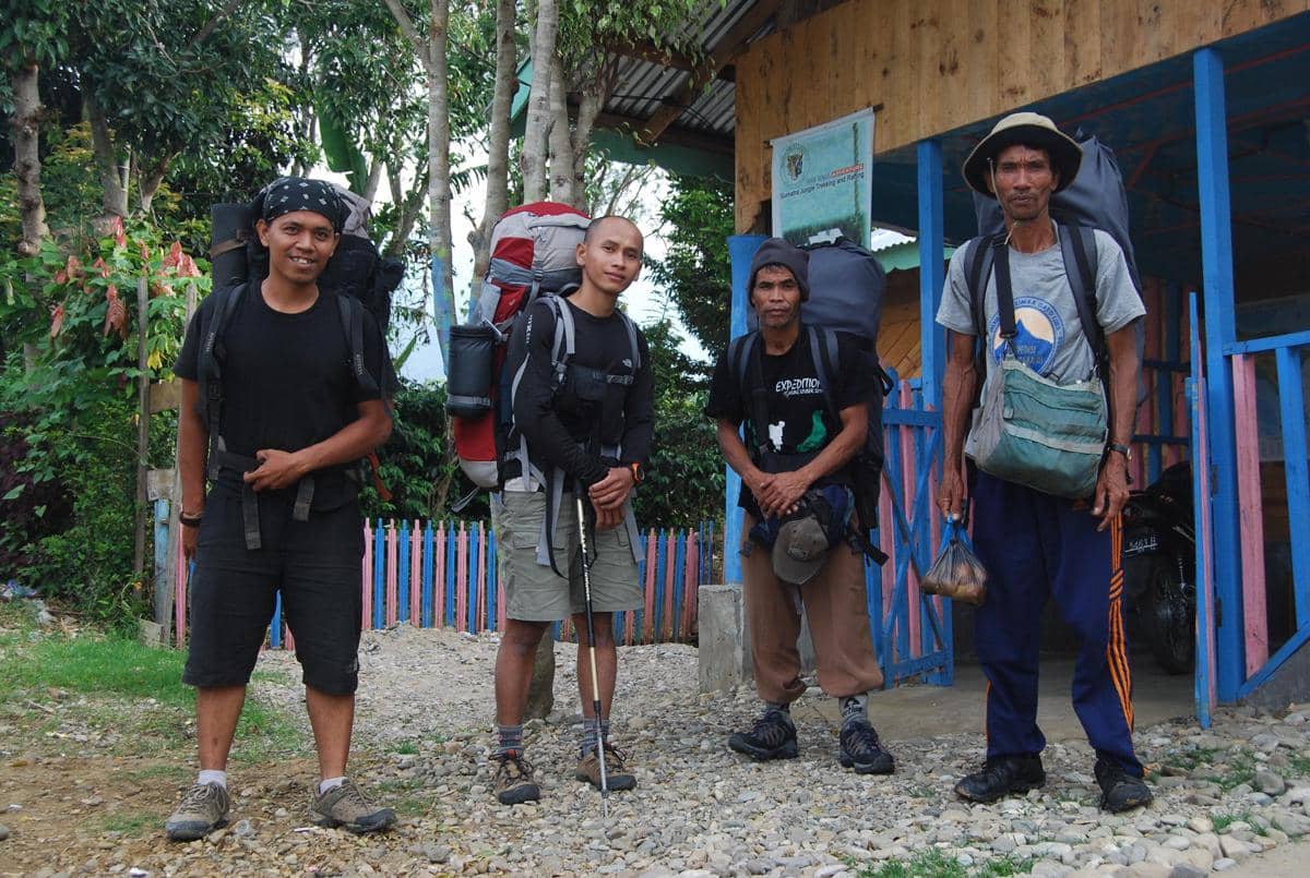 Empat pendaki dengan perlengkapan lengkap berdiri di depan pos pendakian Gunung Leuser yang dikelilingi pepohonan dan bangunan kayu.