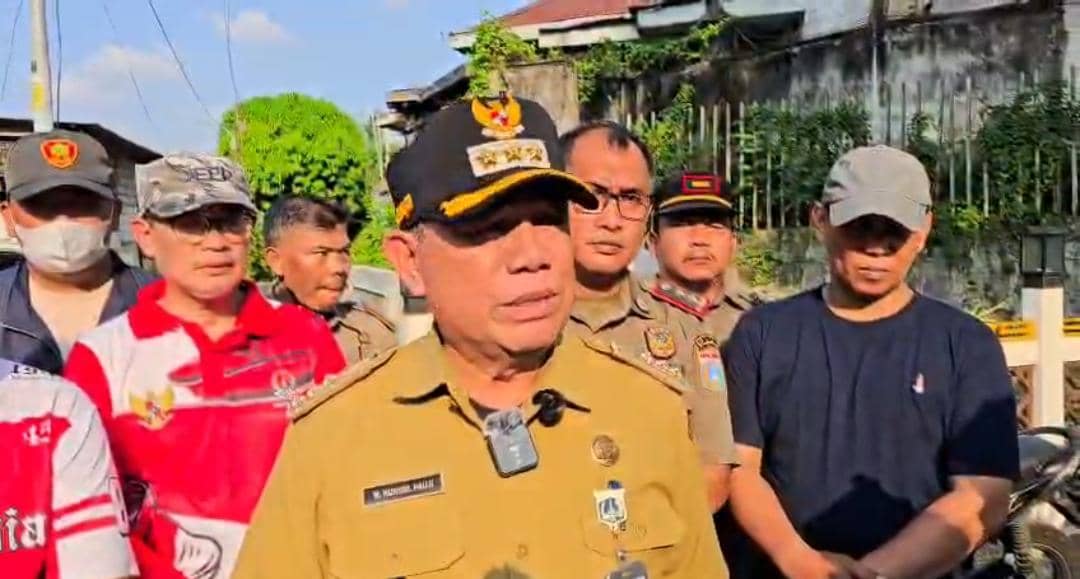 Seorang pejabat kecamatan berseragam cokelat berbicara di luar ruangan didampingi beberapa petugas Satpol PP dan warga di siang hari.