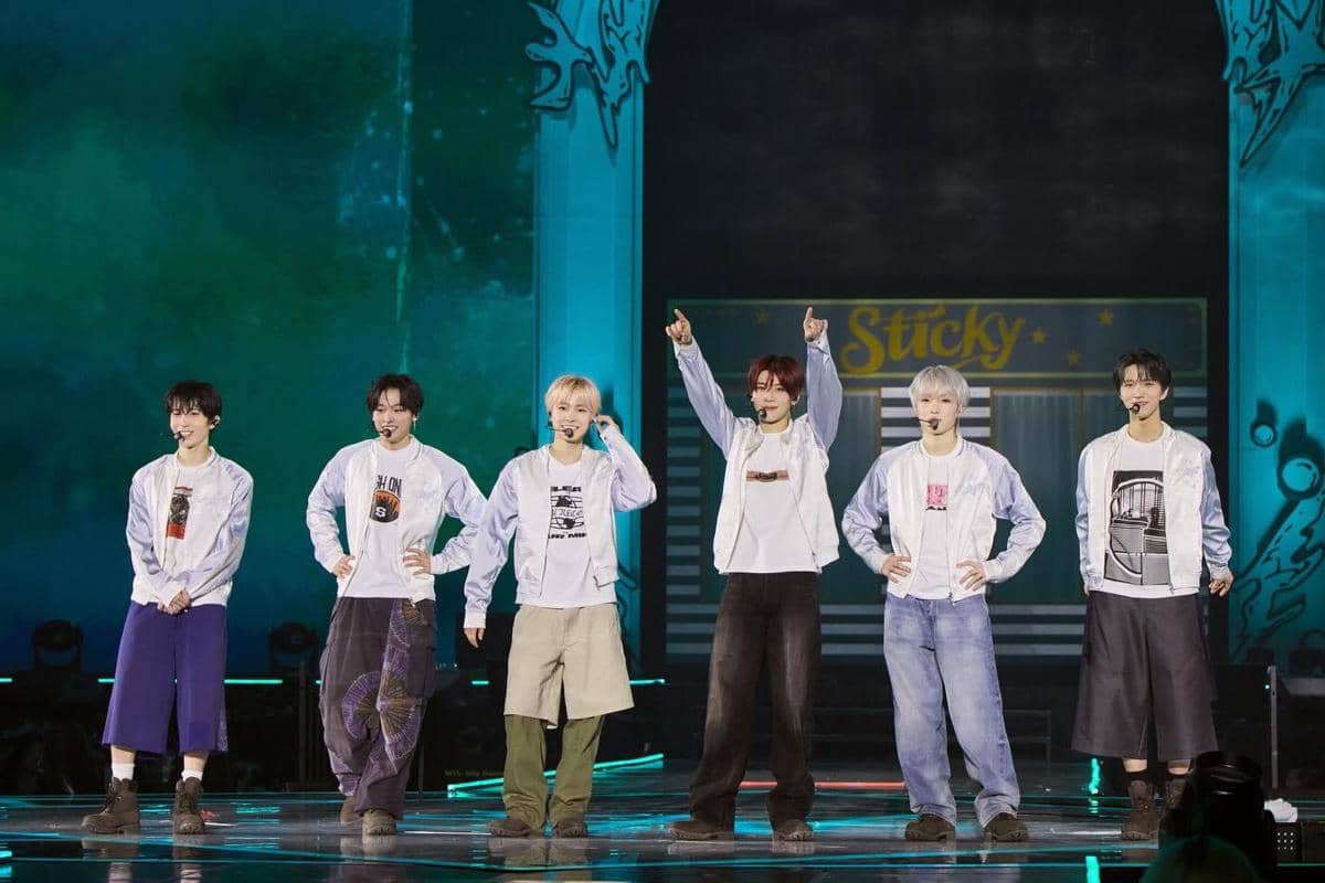 Enam anggota grup musik tampil di atas panggung dengan pencahayaan berwarna hijau dan biru, mengenakan jaket putih dan celana kasual.