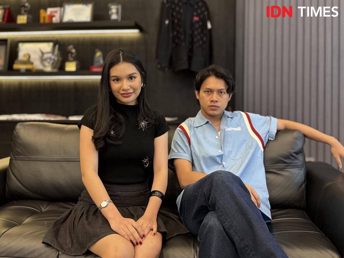 Seorang perempuan dan laki-laki duduk di sofa hitam di ruangan dengan rak berisi piala dan penghargaan di latar belakang.