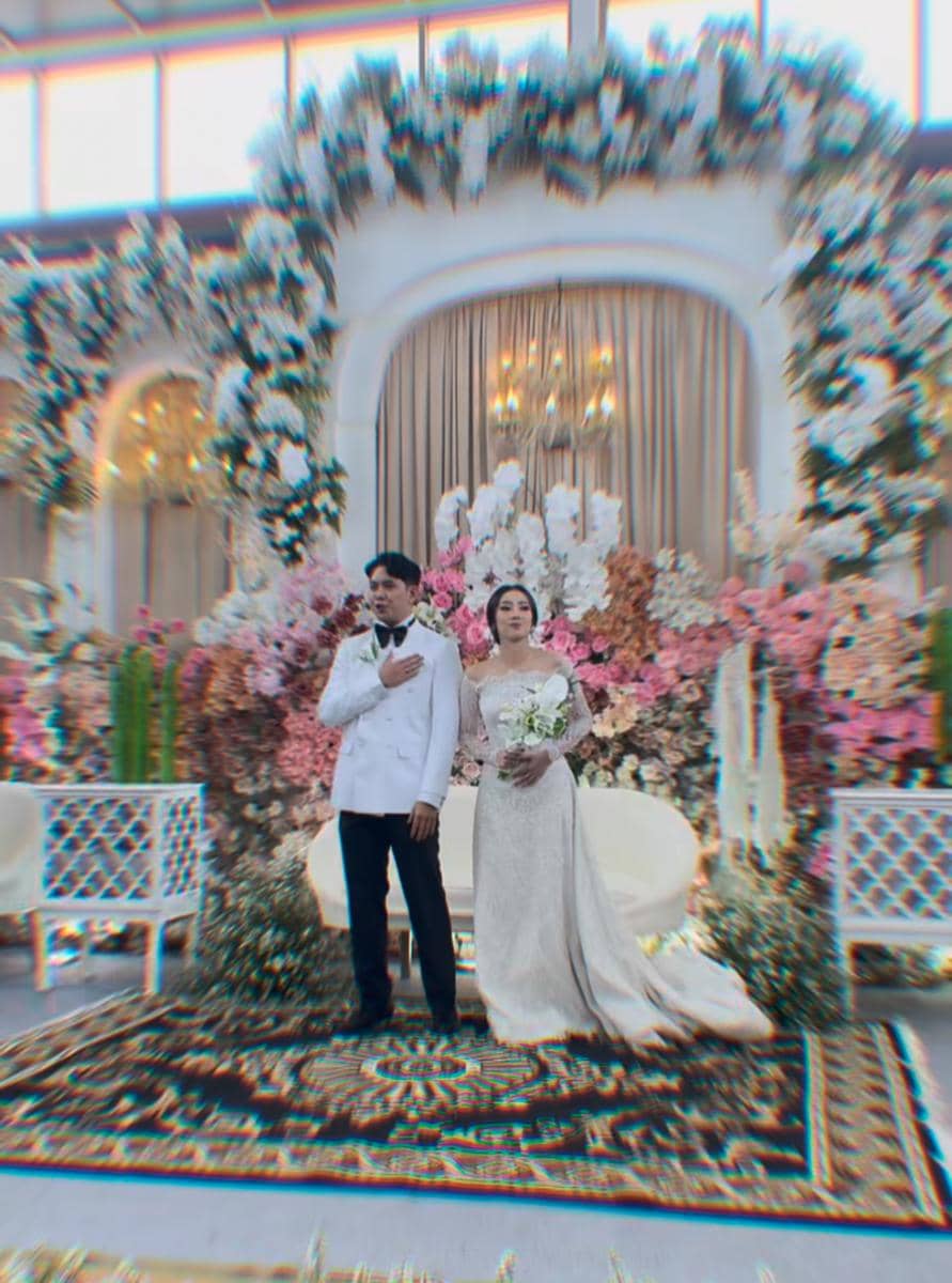 Sepasang pengantin berdiri di pelaminan berhias bunga warna-warni dengan dekorasi mewah dan pencahayaan hangat di sebuah gedung pernikahan.