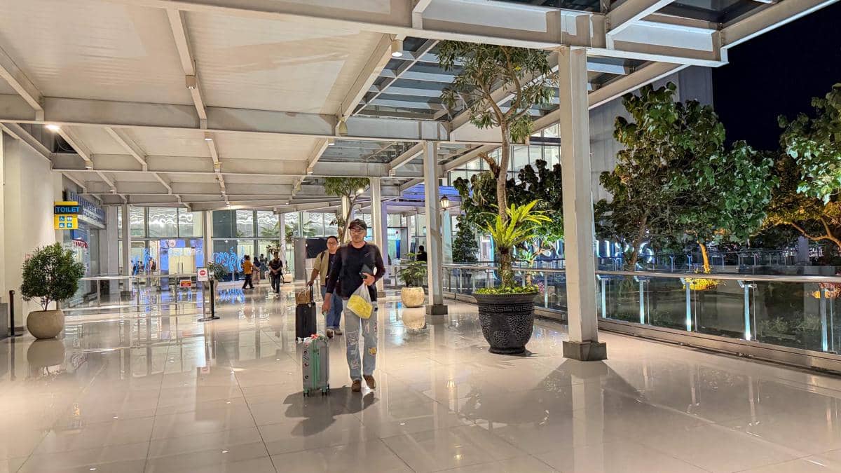 Seorang pria berjalan sambil menarik koper di selasar Bandara Ahmad Yani Semarang pada malam hari dengan pencahayaan terang dan tanaman hias di sekitarnya.