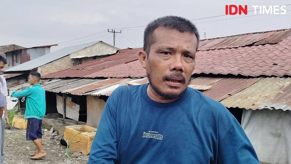 Seorang pria mengenakan kaus biru berdiri di depan deretan rumah beratap seng di kawasan Medan Denai dengan latar beberapa warga lain.