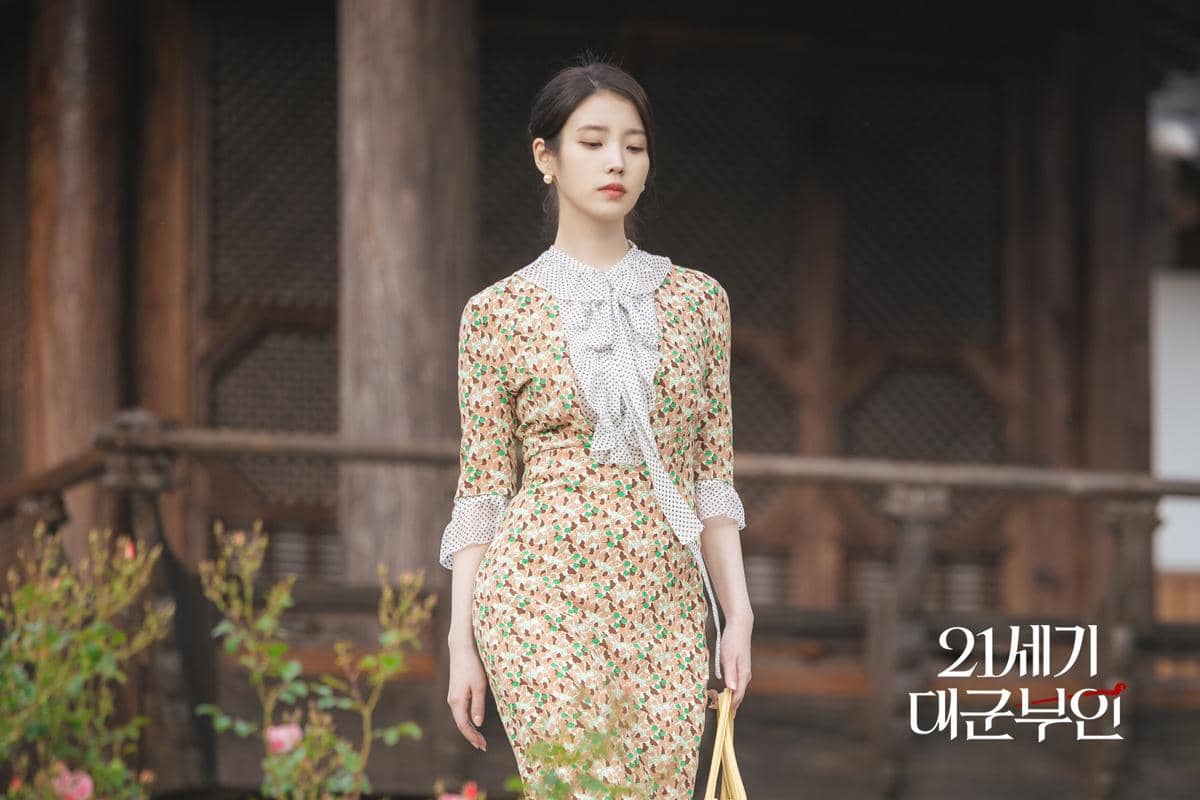 Seorang wanita mengenakan gaun bermotif bunga dengan pita di leher berjalan di depan bangunan kayu tradisional.