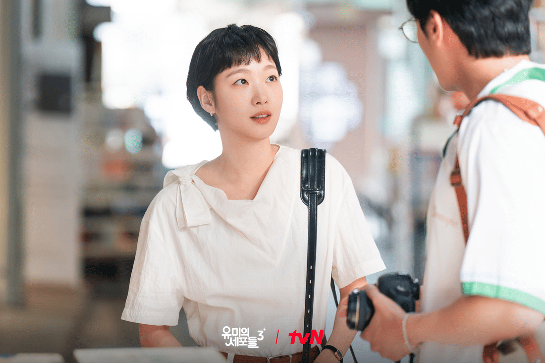 Seorang wanita berambut pendek mengenakan blus putih berbicara dengan pria yang memegang kamera dalam cuplikan drama Yumi's Cells 3.