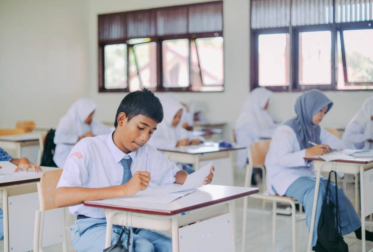 Siswa dan siswi berseragam sekolah sedang fokus mengerjakan tugas di dalam ruang kelas dengan pencahayaan alami dari jendela.