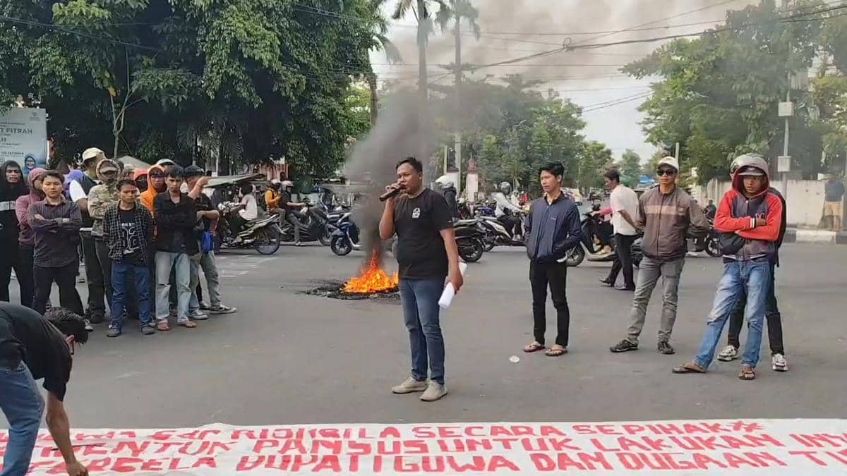 Seorang orator berbicara dengan pengeras suara di tengah aksi unjuk rasa massa Pormula di depan Kantor Bupati Gowa dengan api membakar ban di jalan.