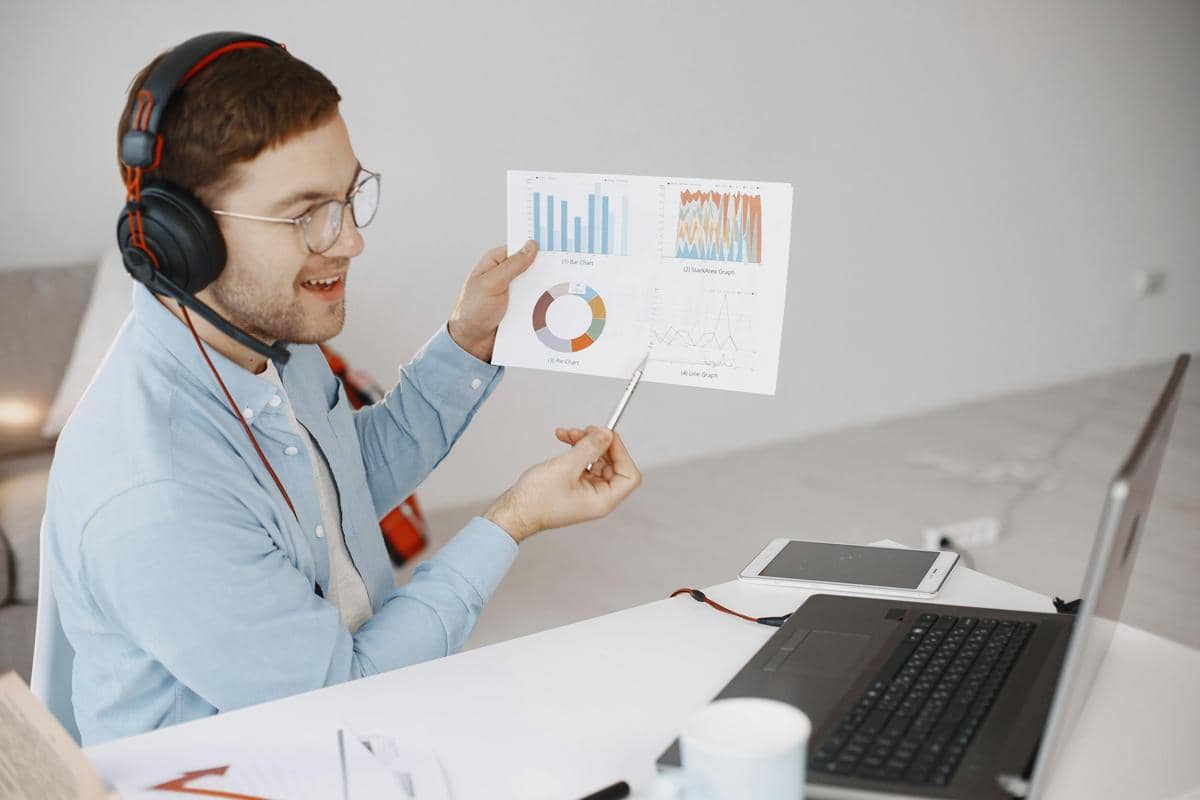 Seorang pria mengenakan headset dan kemeja biru muda sedang menjelaskan grafik laporan kerja di depan laptop di meja kerjanya.