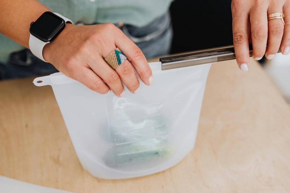 Seseorang menutup kantong plastik ziplock transparan di atas meja kayu dengan kedua tangan, mengenakan jam tangan dan cincin.