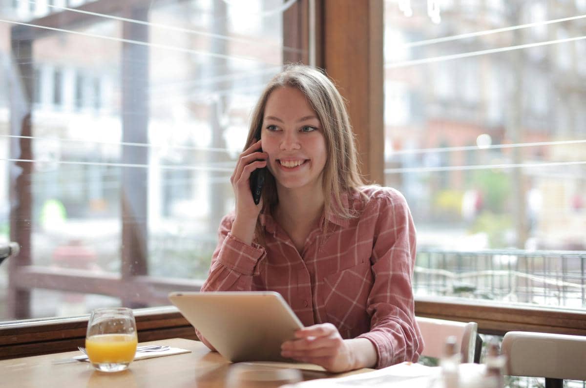 Seorang wanita tersenyum sambil berbicara di telepon dan memegang tablet di meja kafe dengan segelas jus jeruk di depannya.