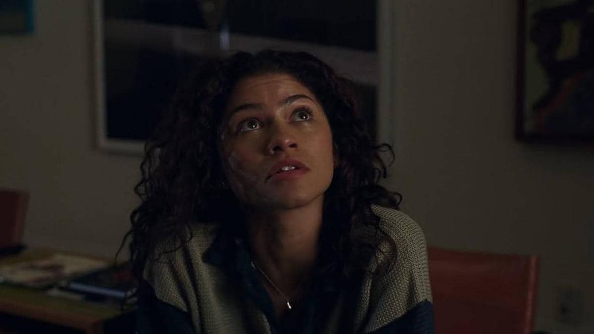 Seorang perempuan berambut keriting duduk di ruangan redup sambil menatap ke atas dengan ekspresi serius dan penuh emosi.