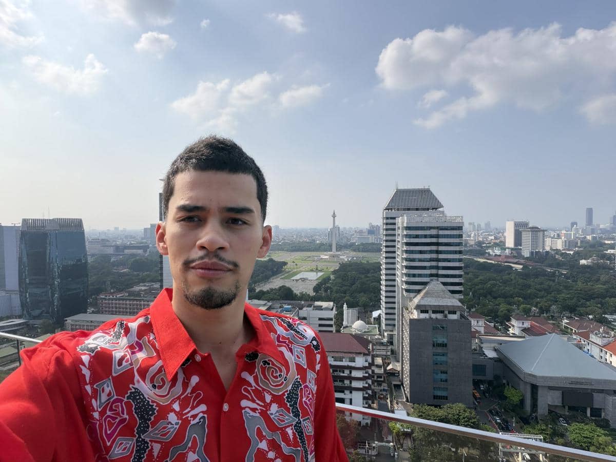 Sneako mengenakan kemeja merah bermotif berdiri di atap gedung dengan latar belakang Monas dan gedung-gedung tinggi di Jakarta.
