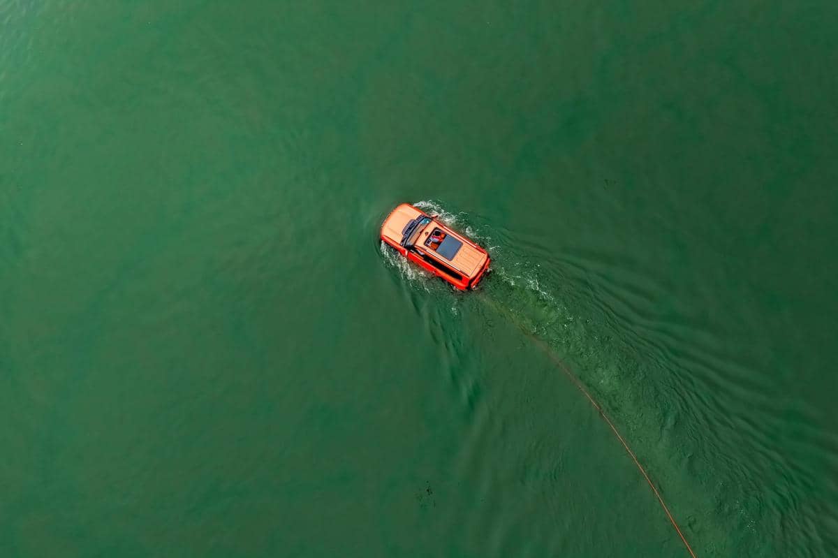 Mobil berwarna oranye melintasi sungai hijau dari pandangan udara, menimbulkan riak air di sekitarnya saat menyeberang.