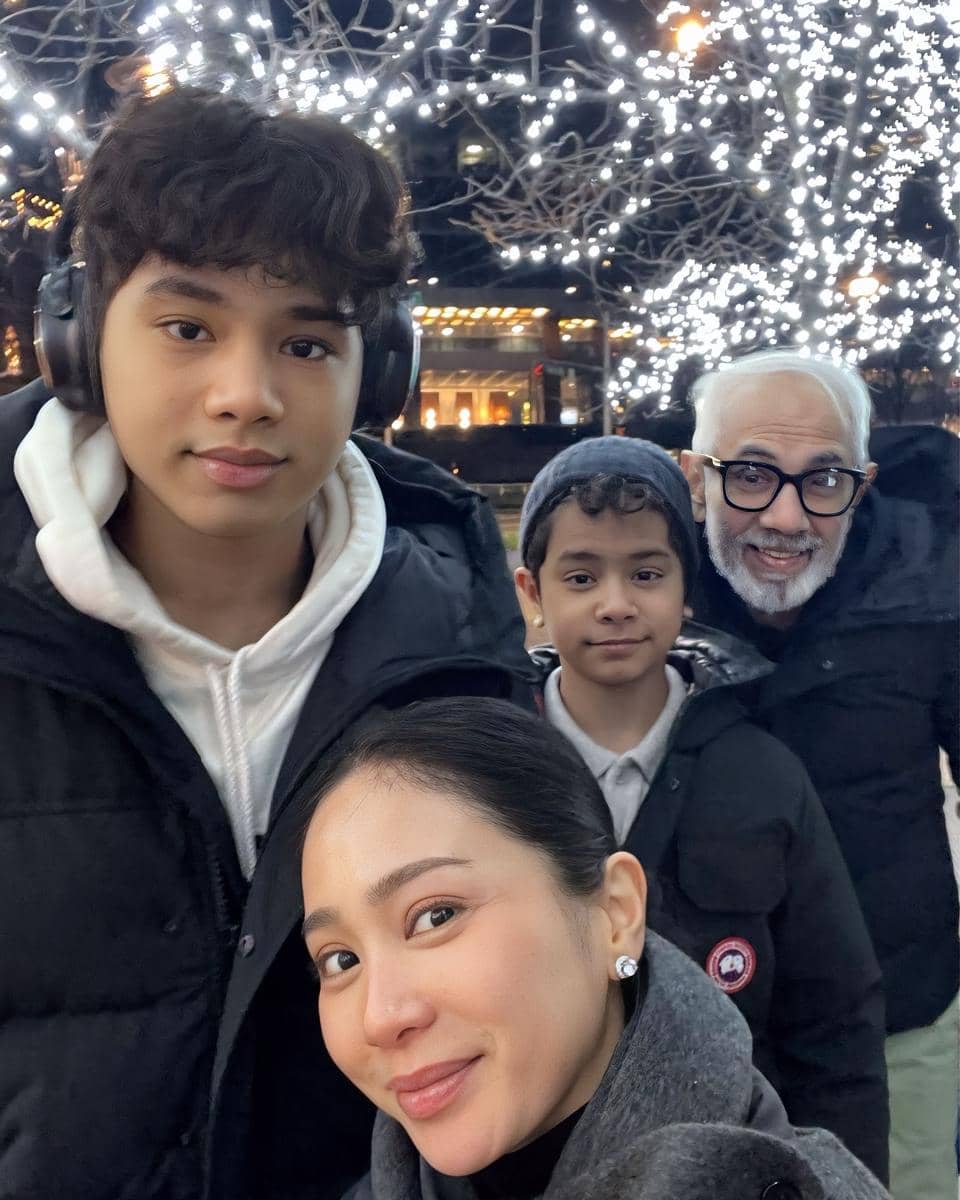 Keluarga Bunga Zainal berpose bersama di malam hari dengan latar pepohonan berhias lampu putih yang berkilauan di area luar ruangan.