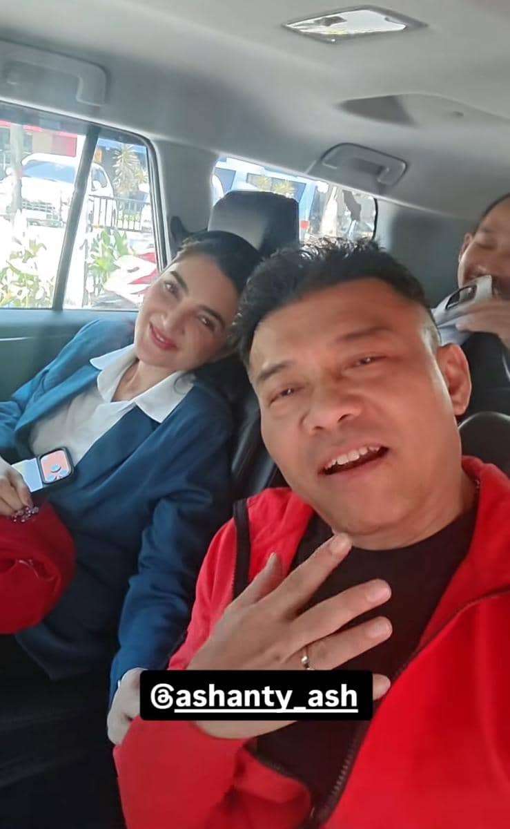 Seorang wanita mengenakan jas biru dan pria berbaju merah duduk di dalam mobil sambil tersenyum ke arah kamera.