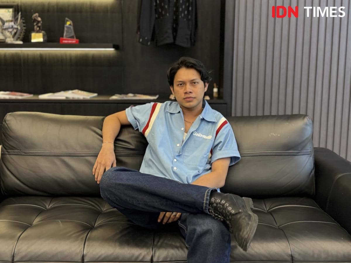 Seorang pria duduk santai di sofa kulit hitam mengenakan kemeja biru muda dan celana jeans di ruangan bergaya modern.