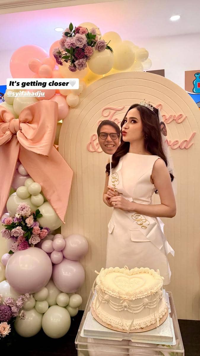 Seorang wanita mengenakan gaun putih dan mahkota kecil berpose di acara bridal shower dengan dekorasi balon pastel dan kue bertema romantis.